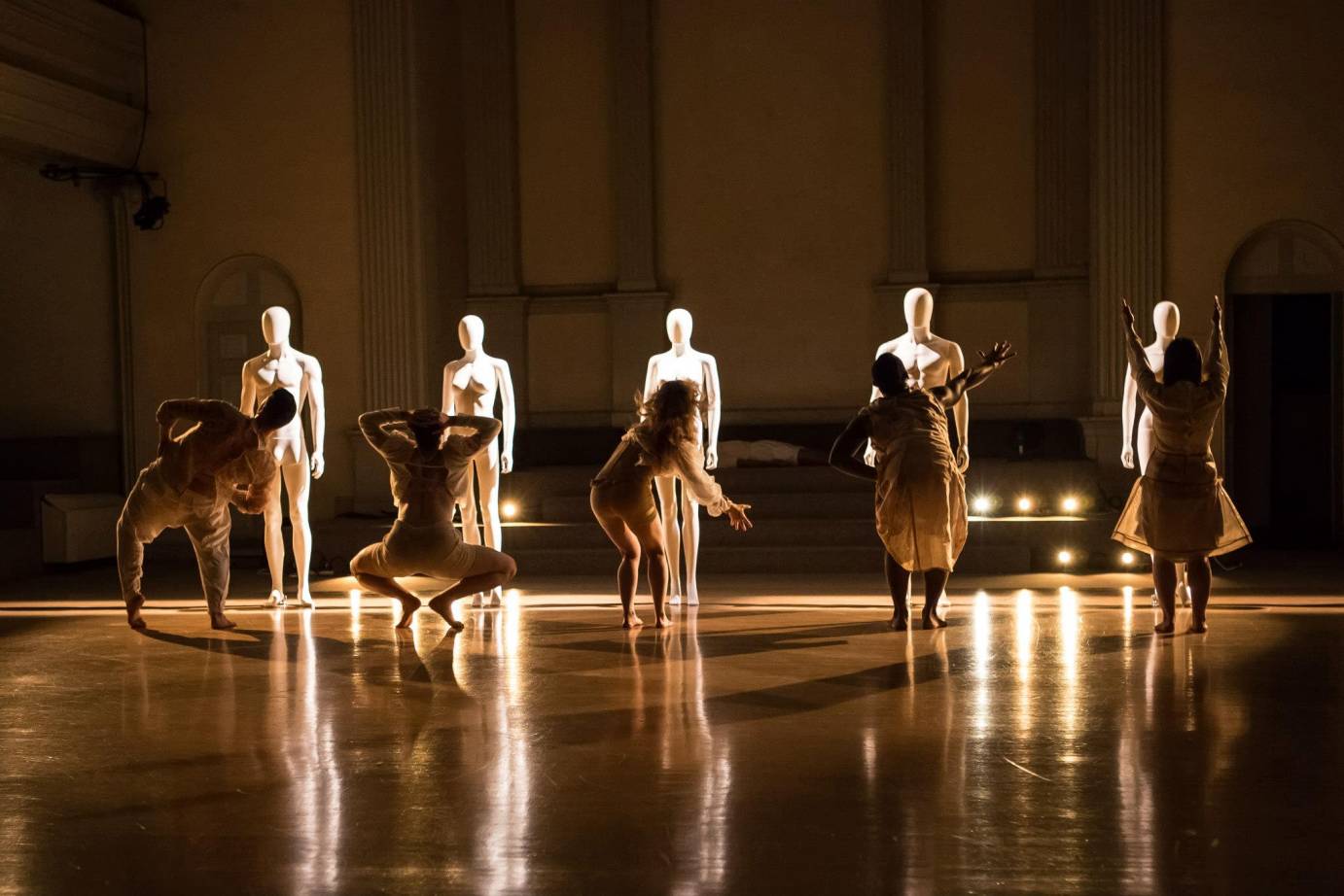 Each dancer gestures to their mannequin counterpart; their backs are towards the audience